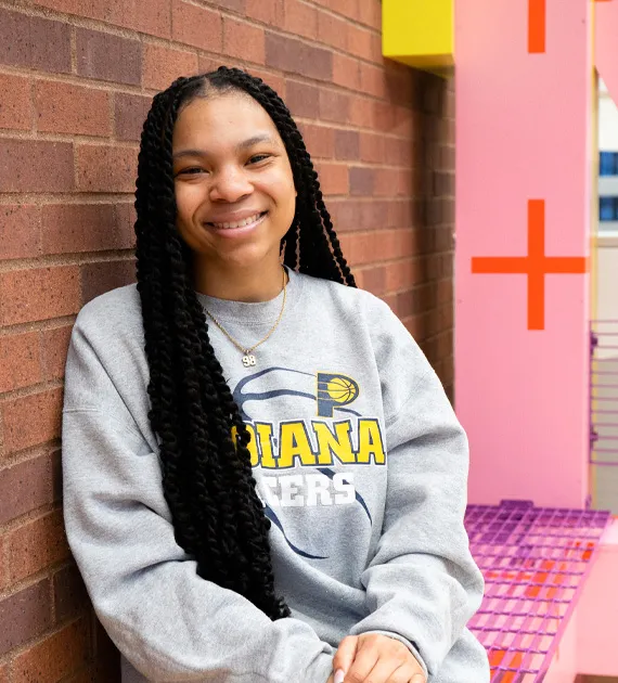 A female student in a gray shirt leaning against a brick wall and smiling. 