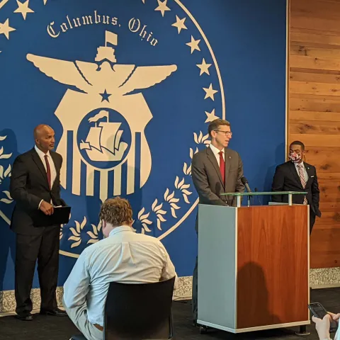 speakers at front of a room at a press conference with reporters