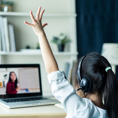 female grade school student raising hand in front of computer with screen showing a teacher