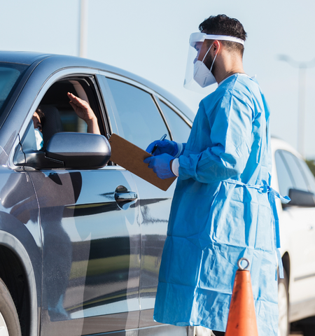 Man in blue scrubs and facemasks taking notes and talking with a person in a car