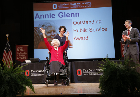 Annie Glenn on a stage in her wheelchair with a person behind her, Dean Trevor Brown to the right, and a PowerPoint image that says "Annie Glenn, Outstanding Public Service Award," behind her