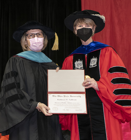 Kathy Sullivan, left, and Ohio State President Kristina Johnson, right, wear masks and academic regalia on a stage holding Sullivan's honorary degree