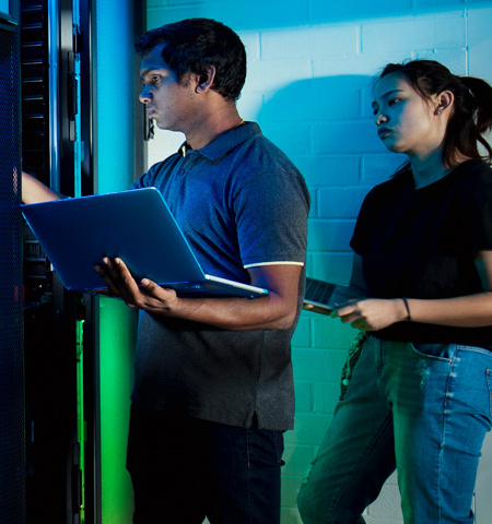 a male and a female holding laptop computers and standing in a computer server room with black, green and blue lighting