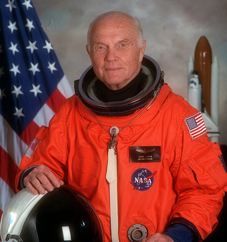 Portrait photo of STS-95 Payload Specialist Glenn wearing the orange partial-pressure launch and entry suit in front of an American flag and space shuttle model replica. 