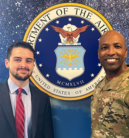 a male civilian and a male Air Force major general standing on either side of an image of the Air Force seal on a wall 