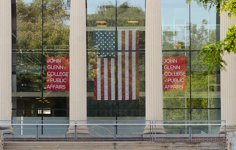 A view of the front window of page hall with two john glenn college of public affairs hanging on both sides of an American flag