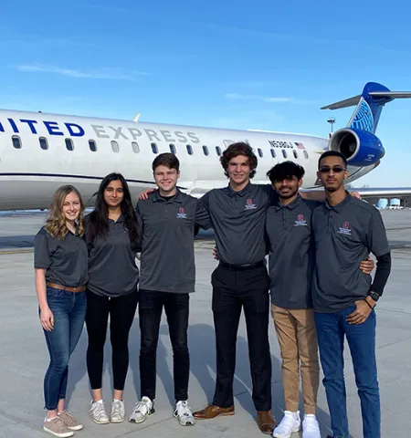 two female and four male students stand in front of a United Express jet at an airport