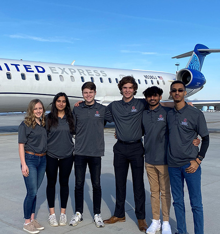 two female and four male students stand in front of a United Express jet at an airport