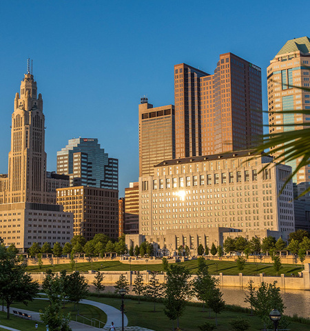 skyline of downtown Columbus during a sunset with a clear blue sky and green foliage on the foreground.