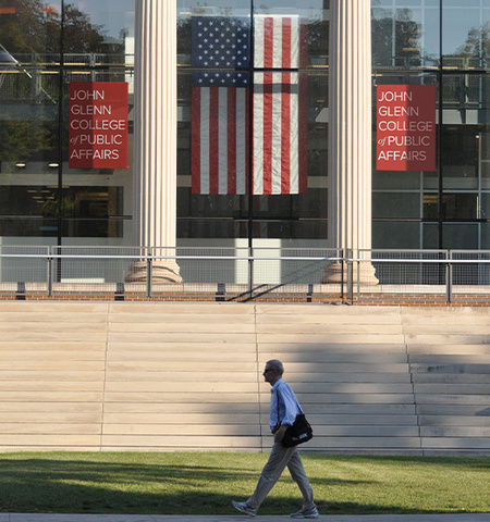 a professional man with a bag over his shoulder walking by page hall's front steps with two john glenn college of public affairs banners and an American flag displayed in the front window.