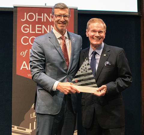 two men standing and holding a triangular award with a John Glenn College of Public Affairs banner in the background