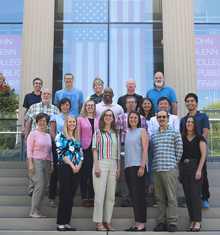 19 men and women stand outside on the steps of Page Hall with windows in the background that have an American flag and two signs that read “John Glenn College of Public Affairs.” 