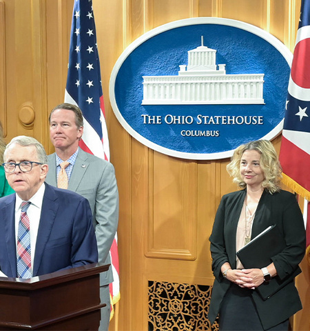 a man standing and speaking at a podium with two women and a man standing behind him. In the background is an oval sign with a white building in it along with the words “The Ohio Statehouse, Columbus” and an American flag and a state of Ohio flag.