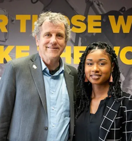 a man and a woman stand inside in front of a wall with the words “for those who make the world move” on it.