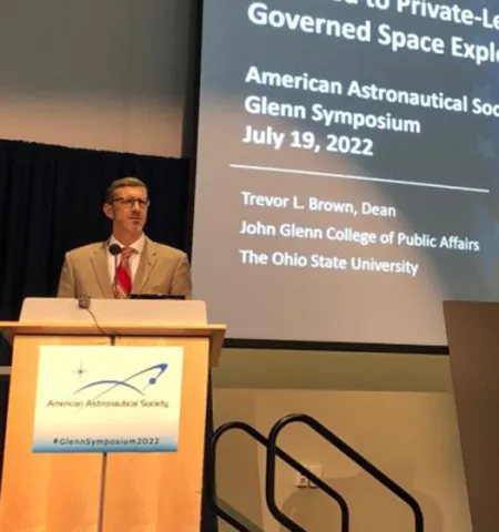 A man stands at a podium with a PowerPoint slide showing behind him