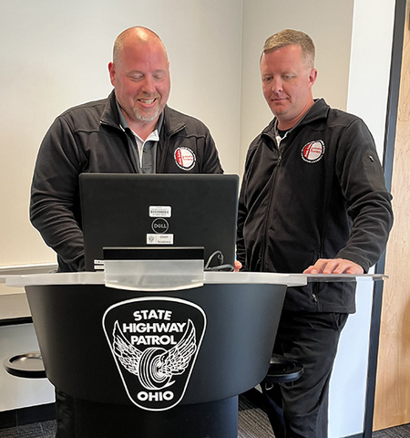 two men stand and discuss leadership principles at a computer at a podium that has a badge in black and white that says “State Highway Patrol Ohio” on the front 