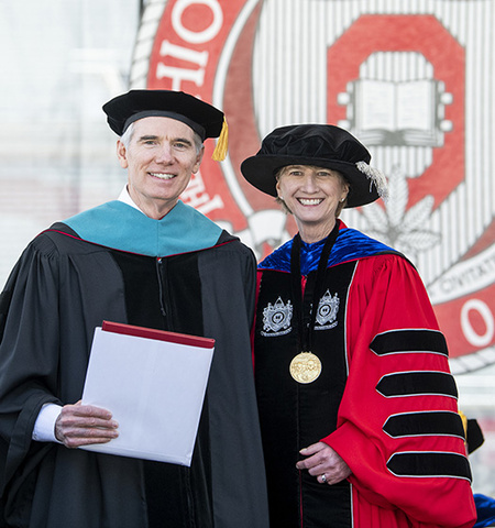 a man and a woman in academic robes and hats stand on a stage with The Ohio State University seal in the background