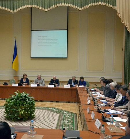 people sitting at tables set in a rectangle in a large assembly hall with a Ukrainian flag along one wall