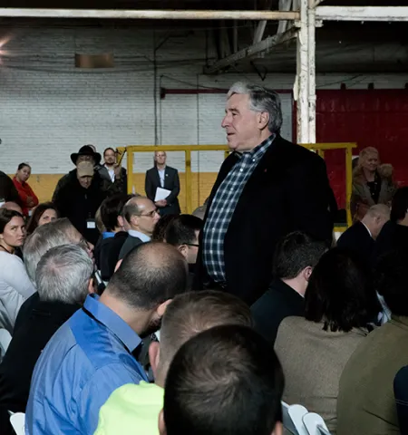 two men stand talking among a seated crowd of people inside a building