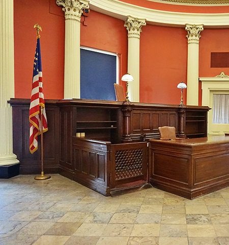 An empty courtroom with a judge bench and witness stand in the middle and an American flag on the left with an orange wall and white pillars in the background (Credit: Robert Linder, Unsplash) 