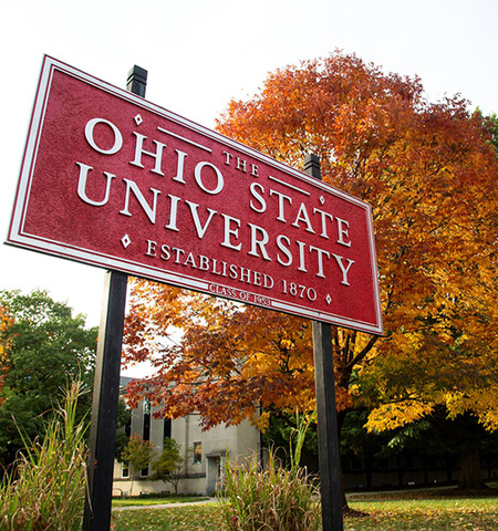 Metal sign with red background and gray letters "Ohio State University" with trees with fall leaves in the background