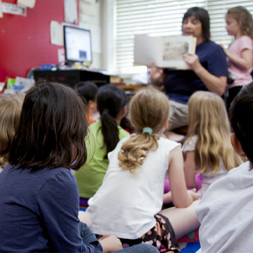 a grouping of young students sitting in front of their teacher and she reads a book to the class.