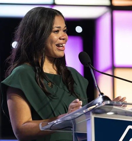 a woman talks at a podium with a microphone and white, purple ad tan blocks of light in the background