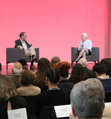 a woman and a man sit on a stage talking with a pink screen in the background and people in a crowd watching in the foreground. A podium on the side of the stage has a sign for the World Food Day summit.