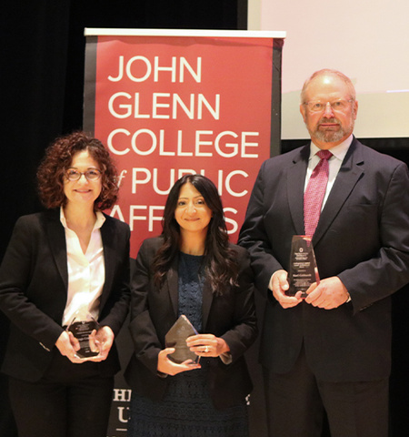 2022 alumni award winners Angie Crandall, Priyanka Bhatt and Karl Gebhardt standing in front of a black and red banner that has the words “John Glenn College of Public Affairs” in white text.