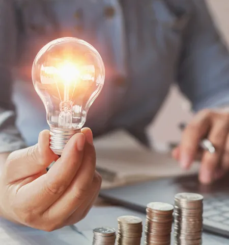 the hand of a person in the background holding a lit lightbulb next to a laptop computer and piles of coins  