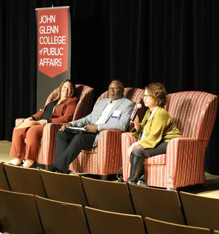a man and two women seated in chairs on a stage talk to an audience.
