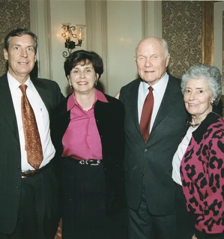 two couples, husbands and wives, stand with their arms around each other and pose for a photo inside a room