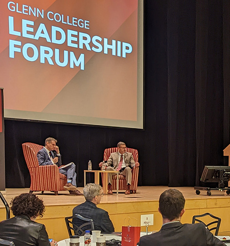 two men sit in chairs on a stage in front of a crowd of people; a screen on the stage reads “Glenn College Leadership Forum”