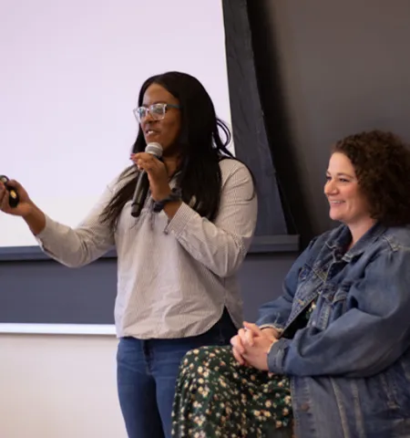 a woman holding a microphone while standing next to a women sitting in a chair.