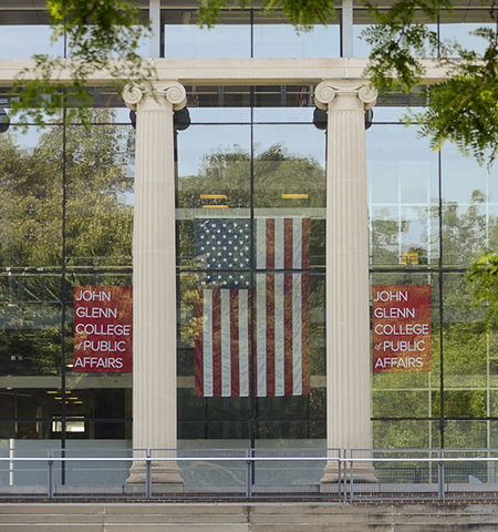 front of Page Hall from outside with columns and, in the windows, an American flag with two signs on either side of the flag that read John Glenn College of Public Affairs