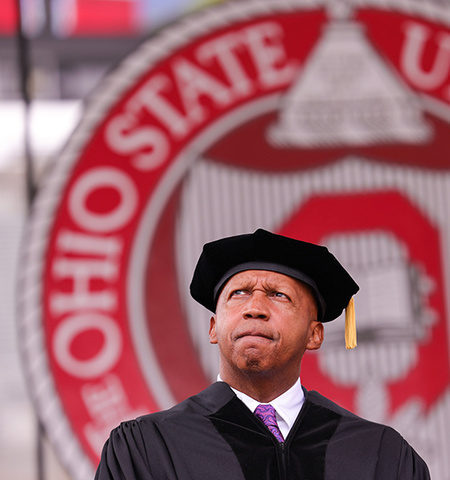 a man wearing academic regalia stands with a thoughtful look in front of The Ohio State University seal