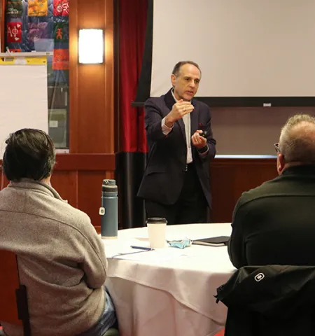 a standing man speaking in front of two men who are sitting.