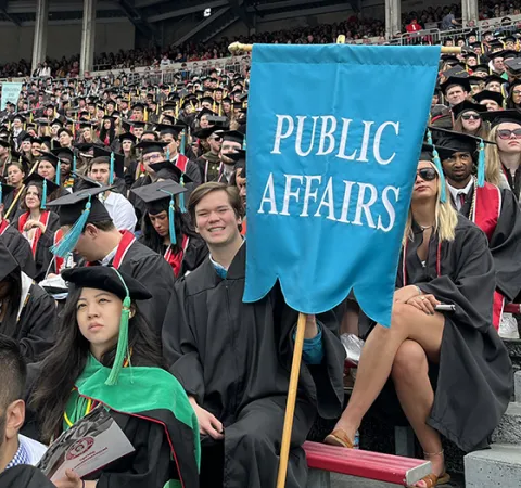 John Glenn students wearing graduation gowns and hats in Ohio Stadium; one male student at the end of a row holds a teal banner that reads “Public Affairs” 