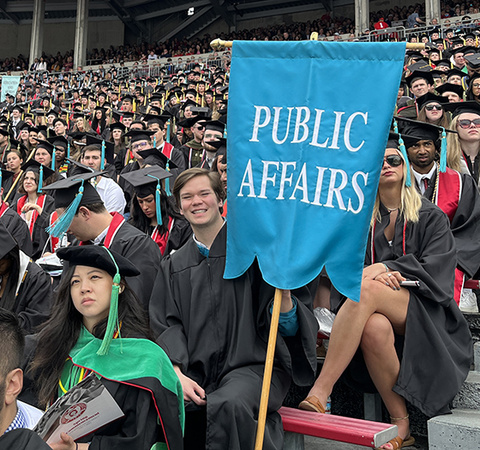 Students wearing graduation gowns and hats in Ohio Stadium; one male student at the end of a row holds a teal banner that reads “Public Affairs” 