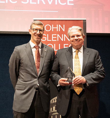 Glenn College Dean Trevor Brown, left, and U.S. Comptroller General Gene Dodaro stand on a stage. Dodaro holds the crystal glass John Glenn Excellence in Public Service award and, in the background, is a partially obscured sign that reads John Glenn College of Public Affairs.