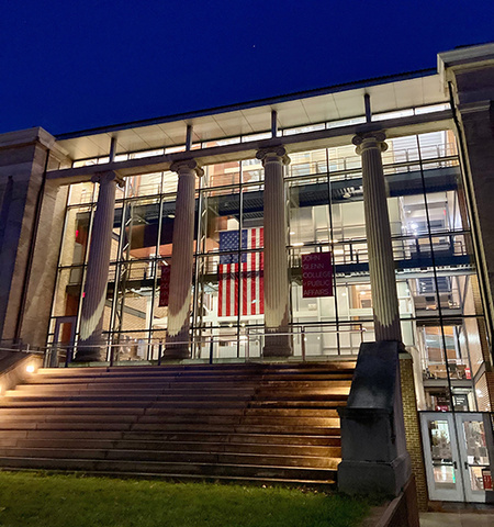 the front of Page Hall lit at night with the American flag and the sign, “John Glenn College of Public Affairs,” in the windows