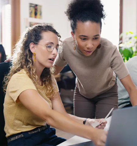 Two women looking at a computer screen in room of other people working.