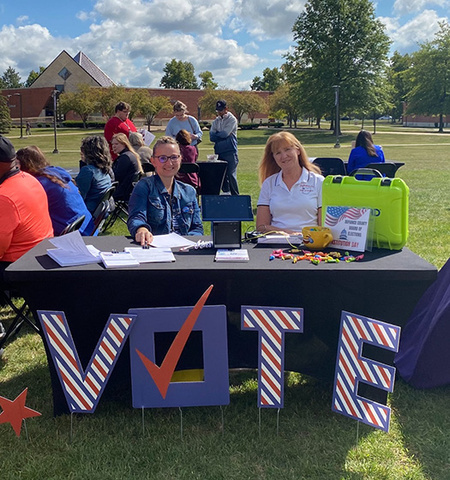  two women outside at an event at a table with a sign that says “VOTE” in front of it. 