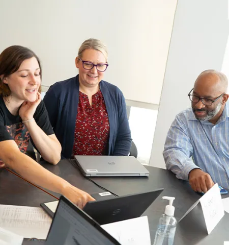 Two women and a man sitting at a table and one women is pointing at a computer screen.