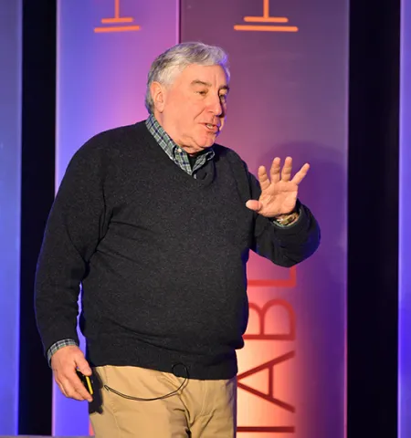 John Glenn College of Public Affairs Professor Ned Hill speaks on a stage with purple and orange panels behind him.