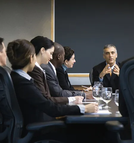 A man at the head of a board table in a meeting room talks to people around the table.