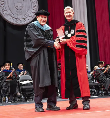 Professor Emeritus Charles Wise, left, upon receiving the University Distinguished Service Award, stands on a stage shaking hands with Ohio State University President Water “Ted” Carter Jr., both in academic gowns.