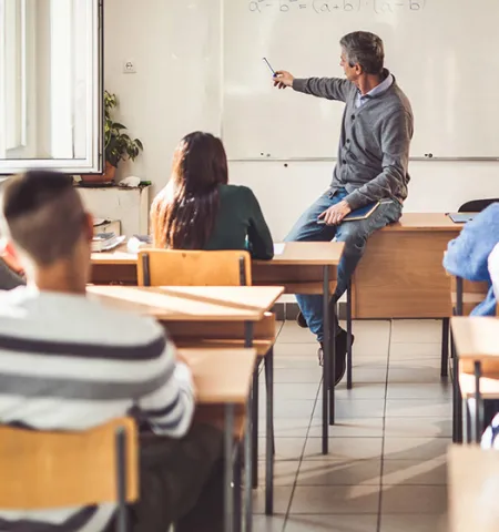 A teacher sits on a desk and points to a white board at the front of students at desks in a classroom