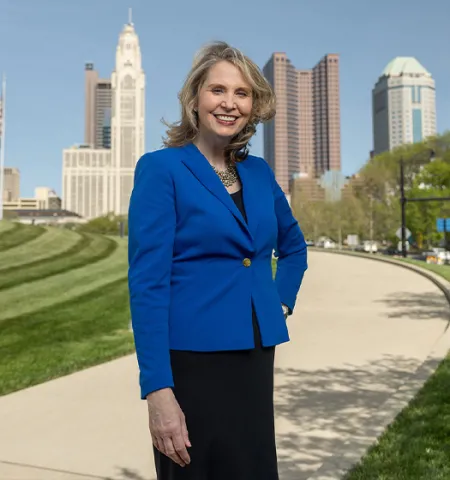 Amy Edwards Taylor smiles and stands with one hand on her hip as she poses for this photo. Behind her is the edge of the Veterans Memorial Museum and farther away, downtown Columbus. She has shoulder length blond hair, wears a business suit and smiles like she's genuinely happy to see you.