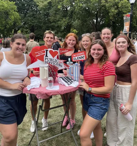 seven students stand around a table outside with tabletop signs celebrating the John Glenn College of Public Affairs